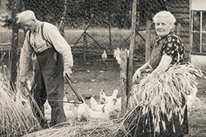 zwart wit beeld met een boer en een boerin met kippen en aan het werk met 					een zeis